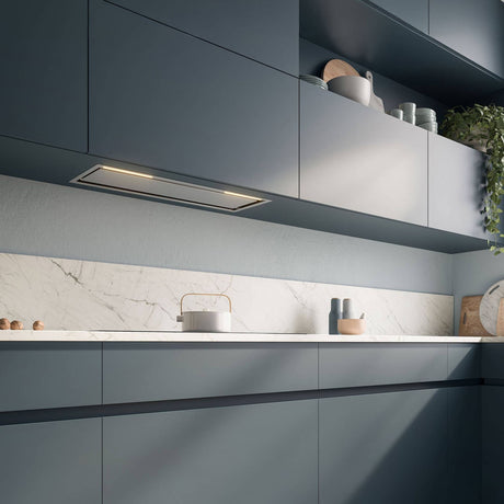Einbauansicht des Falmec Lüfterbausteins Plus Green Tech, bündig integriert in dunkelblaue Oberschränke einer modernen Küche mit heller Marmorarbeitsplatte und Rückwand. Die Dunstabzugshaube ist mit warmweißer LED-Beleuchtung ausgestattet und über einem Induktionskochfeld positioniert.